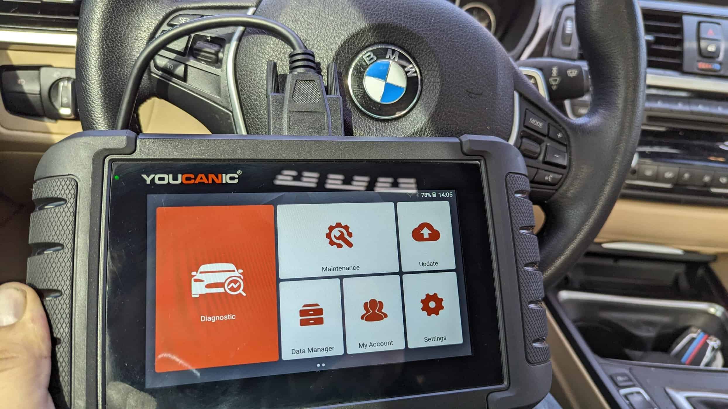 A mechanic holding an OBD-II scan tool while standing at the driver door of a late-model BMW with the hood open, the new battery installed in the trunk
